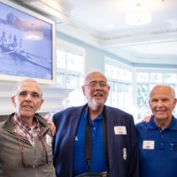 Group of alumni pose together in front of tv displaying a picture of rowing